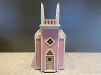 Front view of pink folk art church dollhouse showing four white pointed towers and yellow detailed front door with plastic windows.