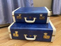 Stacked closed view of two vintage blue hardshell suitcases with gold clasps and white handles.