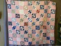 Full view of the vintage patchwork quilt showing the pink squares and multi-patterned blocks arranged in a grid pattern hung vertically against a backdrop.