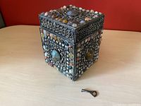 Silver square jewelry box adorned with various polished stones and metal repousse work, showing key in front on wood table