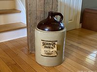 Large vintage Medalta crock jug with dark brown glazed top and gray stoneware bottom with Scarfe Paints label. Shown next to wood box and stairs for size reference.