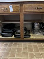 Photo shows inside an open wooden cabinet with multiple stacks of black and grey baking trays and pans on the left side, and large stainless steel pots with lids plus a wire rack on the right side.