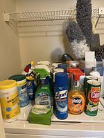 Variety of household cleaning products displayed on a surface including disinfectant wipes, sprays, and cleaning powders. Fluffy cleaning dusters and white plastic bucket visible in background.