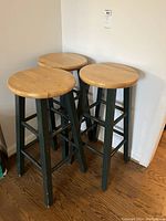 Three wooden stools with light wood round seats and dark legs against a white wall, showing wear and blemishes.
