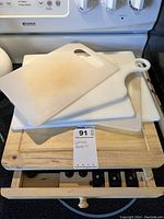 Four white plastic cutting boards stacked on wood channel cutting board with visible stains and marks. Drawer opened to show black kitchen knives and scissors below.