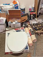 Overall lot arranged on a floor showing the toilet seat on original box, rolled tub mats, plumbing parts, tile mortar and other home improvement materials