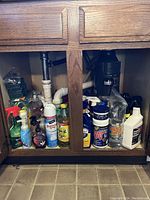 Image of under-kitchen-sink cabinet holding various household cleaning products including spray bottles, pump soap bottles, disinfectants, and insect repellents alongside plumbing pipes.