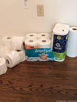 Photo showing Angel Soft 12-pack of toilet paper, a pack of Quilted Northern Ultra Soft paper towels, and some loose toilet paper rolls on a wooden floor.