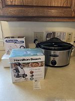 View of Hamilton Beach 2-speed hand blender box, Hamilton Beach oval slow cooker, and Proctor Silex Easy Mix hand mixer box on wooden surface under kitchen cabinets