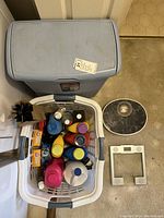 Photo showing laundry basket filled with various detergent bottles, dryer vent cleaning brush, and laundry hamper behind the basket. Two scales are visible on the floor nearby.