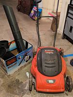 Black & Decker red and black electric mulching mower with corded blower vac parts in box nearby in garage.