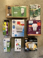Multiple brands and types of boxed and packaged lightbulbs arranged on carpet showing various wattages and features.