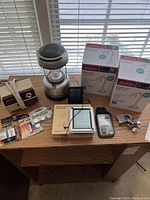 Wide shot of lot contents on a wooden desk near window showing two boxes of Sunbeam lamps, camping lantern, batteries, digital thermometer/hydrometer, pencil sharpener and other miscellaneous items.