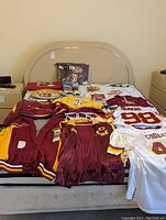 Full view of all items laid out on a bed showing jerseys, jackets, hoodies, shirts, bandana, sunglasses, posters, magnet, ticket, money clip, book, rain poncho, bag with koozies