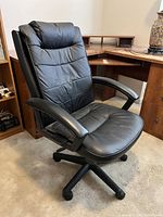 Black cushioned office chair with padded seat and backrest, armrests, five caster wheels, positioned on carpet in front of wooden desk.