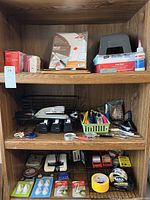 Photo of wooden shelves holding various office supplies including staplers, tape dispensers, scissors, small green basket of pens and clips, calculators, and small office tools