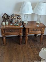 Pair of wood end tables shown with items on top for scale. Each table has a single drawer with metal handle and turned legs, medium brown finish with visible wear and blemishes.