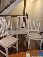 Photo showing the four white armless dining chairs arranged in a cluster, showcasing their vertical slat back design and padded seats with a patterned upholstery.