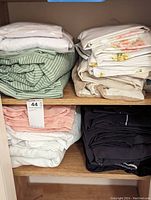 Photo showing piles of various folded bed linens and towels arranged on shelving, including green striped linens, floral linens, beige, white, peach, and black linens.