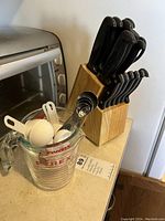 Photo of a wooden knife block with black-handled knives and a Pyrex glass measuring cup holding white plastic measuring spoons