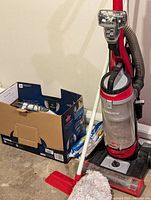 Red and black BISSELL Cleanview upright vacuum cleaner with clear dust canister, standing next to a box containing vacuum bags and cleaning supplies, plus a mop base and soft cloth on floor.