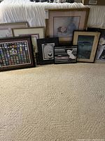 Wide shot of nine framed wall art pieces on carpet, including floral prints, landscape, Patrick Nagel style portraits, Super Bowl 50 poster, and a 1972 framed poster.