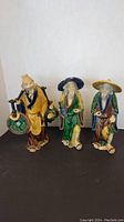 Three Asian ceramic statues: one water carrier and two fishermen, all approximately 10 inches tall, displayed side by side on a dark surface with a plain background.