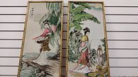 Photo showing two gold bamboo-style framed silk tapestries hanging side by side on a wall with white horizontal paneling. The tapestries depict two different Asian ladies in traditional clothing with detailed natural surroundings.