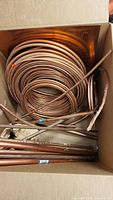 Copper pipe coils tightly rolled placed inside a box showing multiple diameters and some straight pipe pieces.