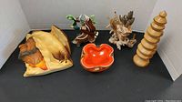 Group photo of lot contents showing wooden carved mini tree, ceramic flying duck, ceramic Middle Eastern man with hawk, shell pickle fork holder, and retro amber-orange glass bowl.