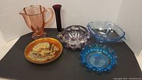 All six items in the lot including the amber pitcher, amethyst vase, amber lid dish, purple rounded glass bowl, blue divided dish, and bright blue ruffled bowl.