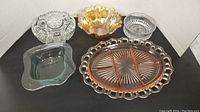 Five glass pieces on dark surface including opalescent orange candy bowl, cranberry tray, cut glass bowl, iridescent scalloped bowl and large decorative tray.