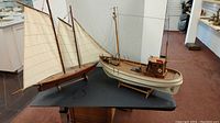 Two wooden model boats displayed on a black table, one sailboat with three sails, and one motor boat with cabin and mast.