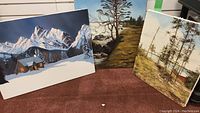 Three acrylic canvas paintings by Irmie Pauls displayed on carpet. Winter mountain cabin, shoreline tree scene, and pine trees by shore.