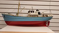 Side view of wooden model trawler showing red hull, light blue upper hull, white superstructure, yellow mast.