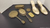 Photo showing all items in the lot laid out: pair of vintage shoes, three-piece gold-tone vanity brush set, compact case, and medallion.