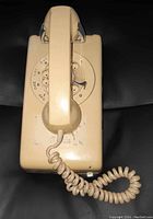 Beige rotary dial wall phone with handset shown from front. Visible signs of use and discoloration. Coiled cord attached to handset.