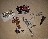 All seven brooches laid out on a textured surface showing each piece: JJ Jonette pewter frog, gold-tone frog with red rhinestones, three pig ballerinas, man rolling dice, rhinestone scissor brooch, straight razor signed Weck Hair Shaper, and plastic cat pin.