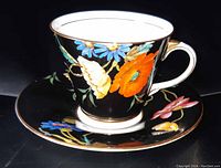 Angled view of Aynsley cup on saucer with vibrant floral hand painting on black background, gold trim visible