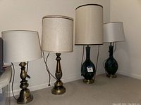 Four lamps side by side on floor; two brass base lamps with off-white shades and two green glass base lamps with taller off-white shades.