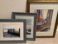 Three framed prints leaning against wall, front views showing beach scenes and bathtub scene
