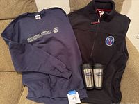 Photo showing navy blue Centennial Airport sweatshirt, black/navy fleece vest, and two branded coffee cups on a beige chair.