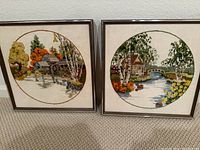 Two framed crewel embroidery wall hangings side by side on floor showing detailed scenic needlework in circular designs.