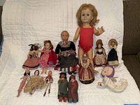 Full collection of 14 vintage dolls arranged on white quilt backdrop showing diversity in sizes and styles
