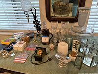 Photo showing assortment of tableware including goblets, vases, candles, decorative items, and small coasters on a table under window blinds, with large framed vintage mirror in background