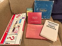Photo showing six vintage game and book items on a fabric surface: Monopoly, two Trivial Pursuit boxes, Scrabble, Official Scrabble Players Dictionary, and The Prophet by Kahlil Gibran.