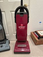 Front view of maroon/red Hoover Sprint upright vacuum cleaner with black hose wrapped at the top, standing against door and carpeted floor.