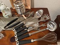 Close-up of metal cooking utensils including slotted spoon, pasta server, ladle, whisk, and tongs with black handles laid out on dark wooden surface