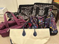 Photo of all bags: three black and white geometric pattern bags (rolling suitcase, tote bag, small bag), maroon duffel, floral duffel, and cream tote with blue handles.