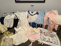 Vintage womens and baby clothing laid out on a floor against a wall, including gowns, dresses, bonnets, and a colorful scarf.